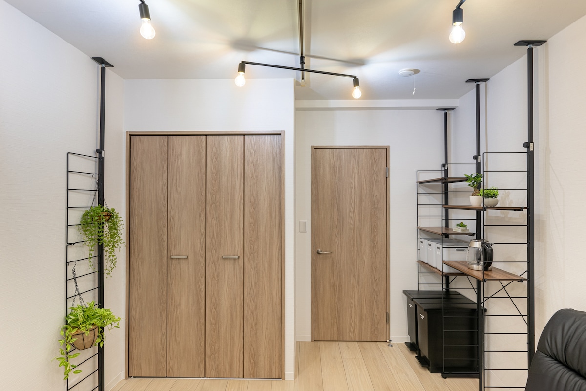 An organized workspace is featured, showcasing wooden cabinets with sleek handles and a door on opposite sides. Industrial-style shelving holds various plants alongside storage boxes, creating a functional and minimalist environment. Soft lighting from overhead fixtures enhances the clean, modern design.