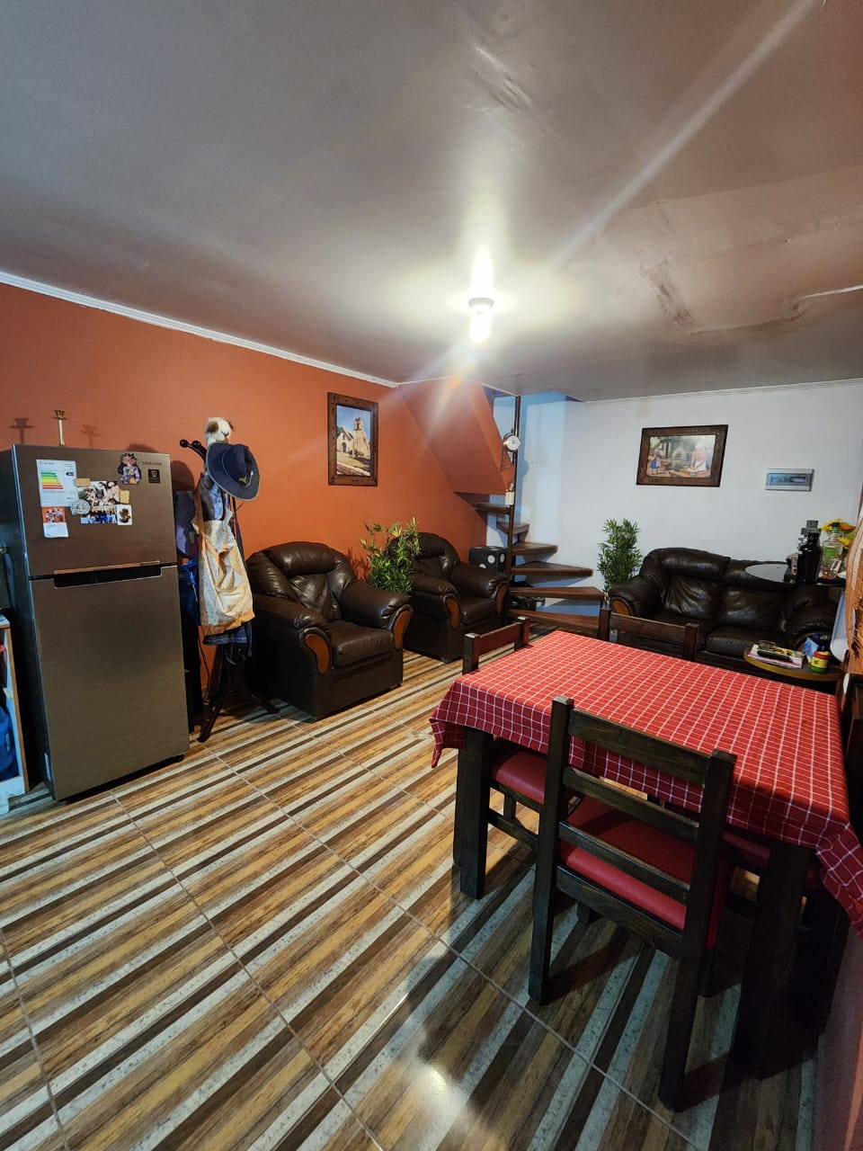 A compact living area features brown leather seating and a red-checkered dining table set for four. A refrigerator stands against one wall, while indoor plants add a touch of greenery. A staircase is visible in the background, leading to another level.