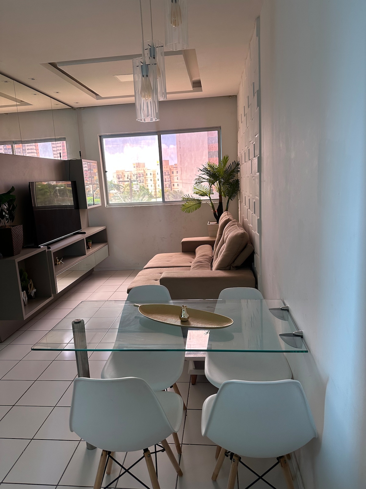 An open living space features a glass dining table surrounded by four white chairs. A light beige couch is positioned near a large window, providing natural light. A minimalist bookshelf and a television unit are present against one wall, with green plants adding a touch of nature.