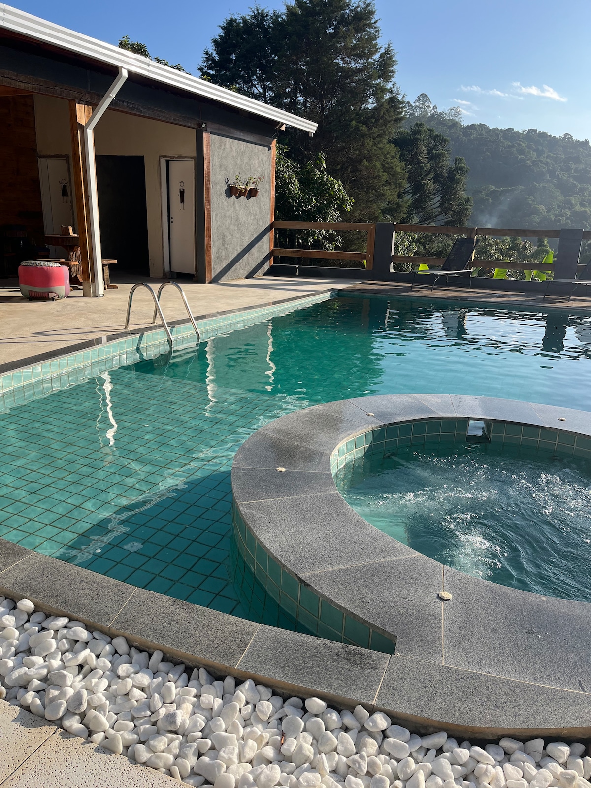 An inviting swimming pool features a circular hot tub, with smooth stone edges and clear blue tiles. Sunlight reflects off the water's surface, enhancing the serene atmosphere. The surrounding deck is made of wood, seamlessly connecting to a cozy outdoor space.