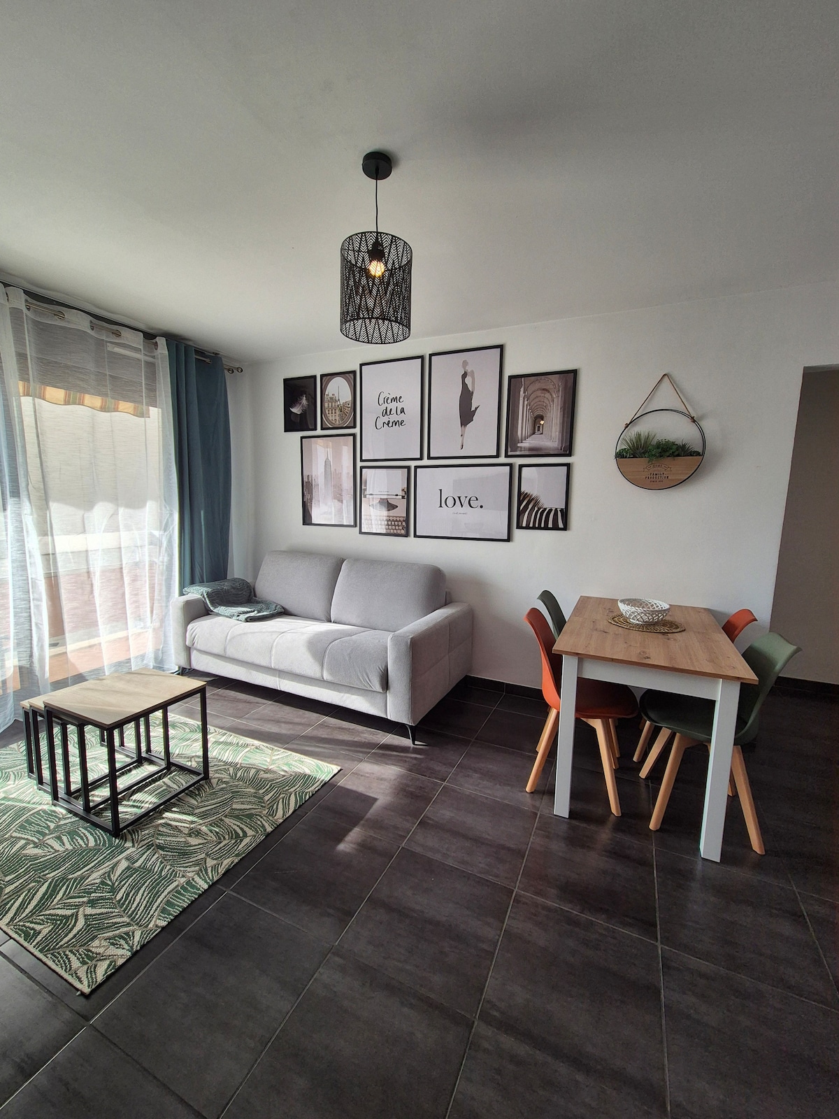 A living area is furnished with a convertible gray sofa and a wooden dining table for four, surrounded by chairs in various colors. A patterned rug adds texture to the floor. Wall art features diverse black-and-white photographs, enhancing the room's ambiance.
