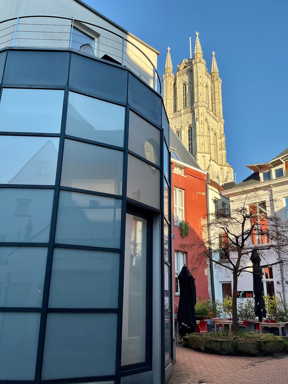 A modern glass structure is positioned among charming buildings, with a tall historic church spire visible in the background. The scene is set against a clear blue sky, highlighting the contrast between contemporary architecture and traditional elements.