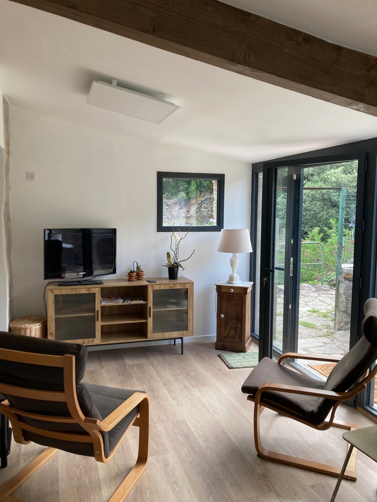 A bright living area features a flat-screen television mounted on the wall, with a wooden cabinet and comfortable seating. Large glass doors open to the outside, allowing natural light to fill the space. A small lamp and decorative elements add a touch of warmth.