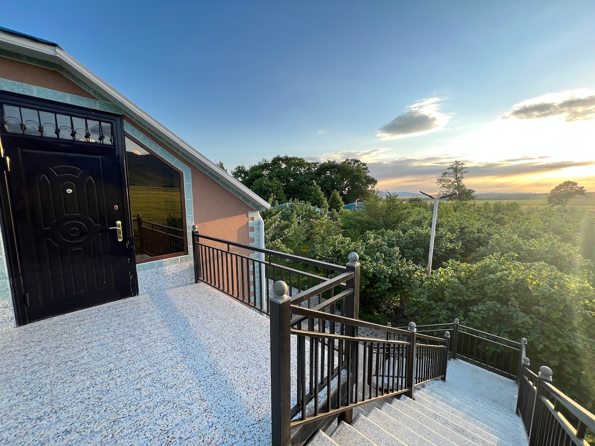 A well-maintained staircase leads to a black front door, surrounded by a verdant landscape. Sunlight casts a warm glow over the scene, highlighting the natural greenery in the background, while a partially cloudy sky adds depth to the horizon.