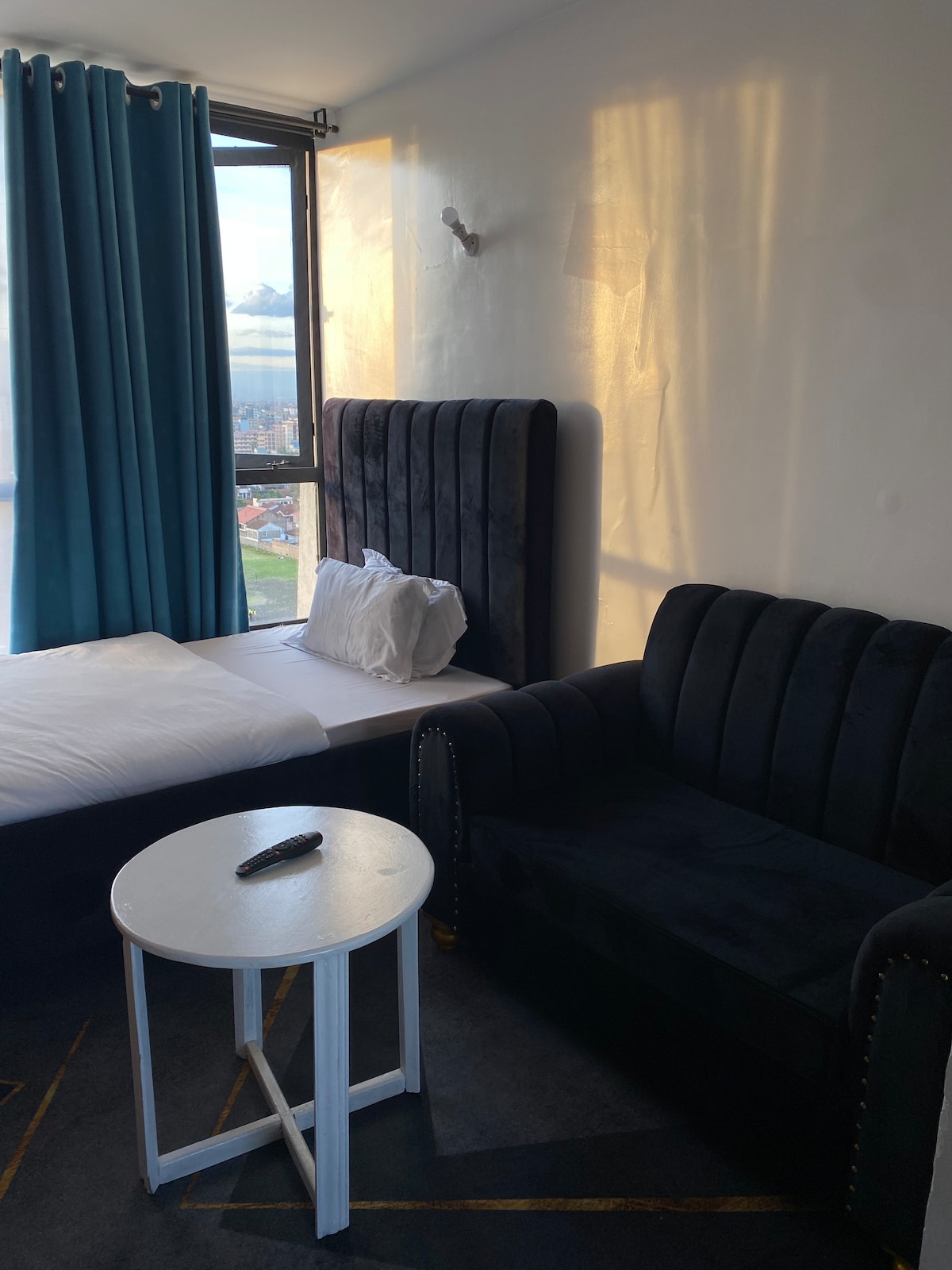 A cozy sleeping area features a spacious bed with a white duvet and a plush black upholstered headboard. A black sofa sits beside a small white table. Natural light enters through a window covered by floor-length blue curtains.