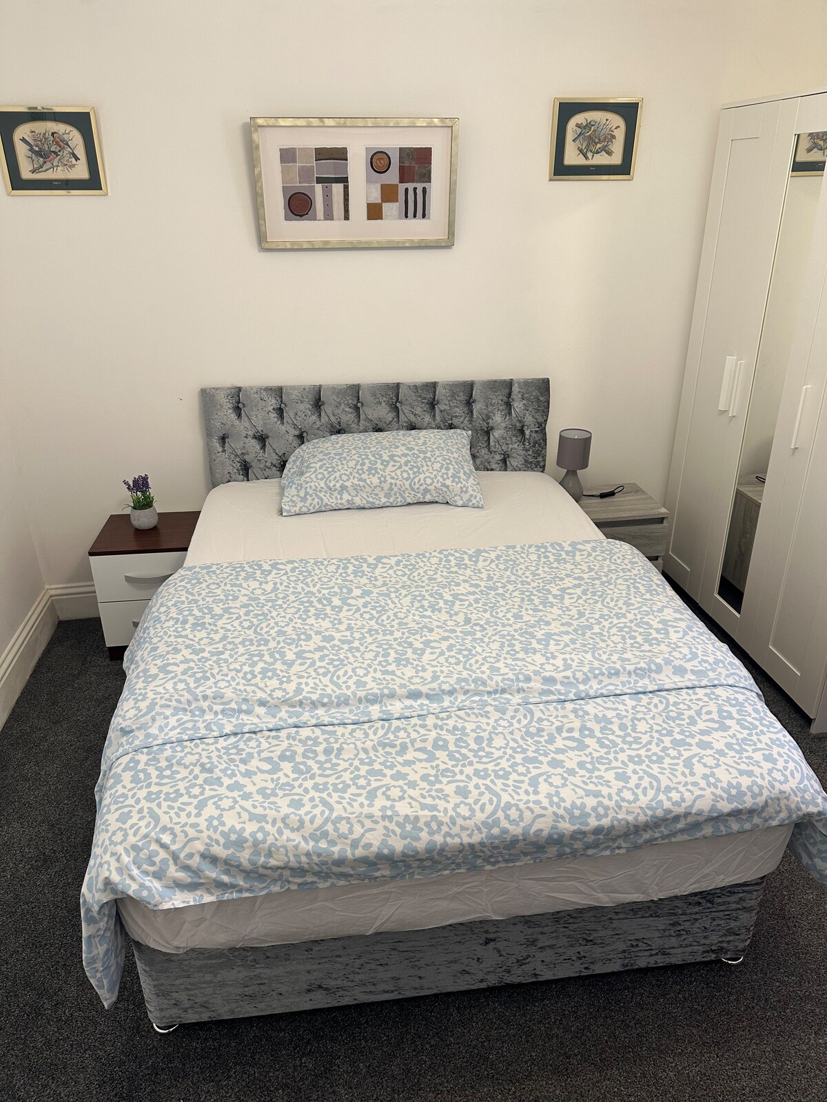 A neatly arranged bedroom features a bed with a light blue and white patterned coverlet. A small bedside table with a lamp and stationery is positioned next to the bed. A white wardrobe stands against the wall, accompanied by framed artwork above the headboard.