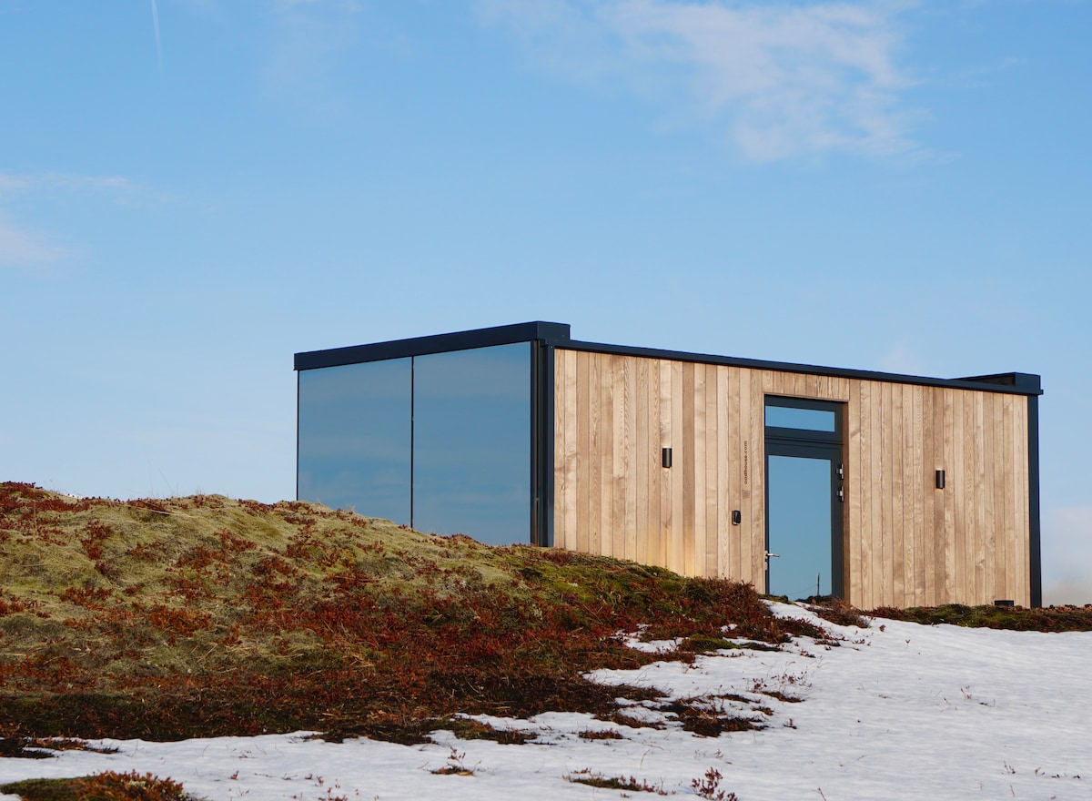 A modern mirrored house is positioned on a gently sloping landscape. Its wooden exterior contrasts with the surrounding snow and earthy tones. Large glass panels reflect the sky, while the entrance is framed by a sleek door and wall-mounted lights, enhancing its contemporary design.