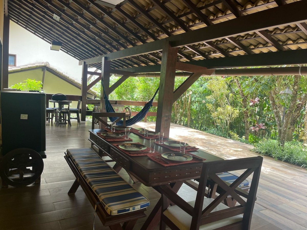An outdoor dining area features a long wooden table set with plates, surrounded by a spacious deck. Hammocks are suspended nearby, and lush greenery provides a natural backdrop. The sloped roof adds architectural interest while offering shade.
