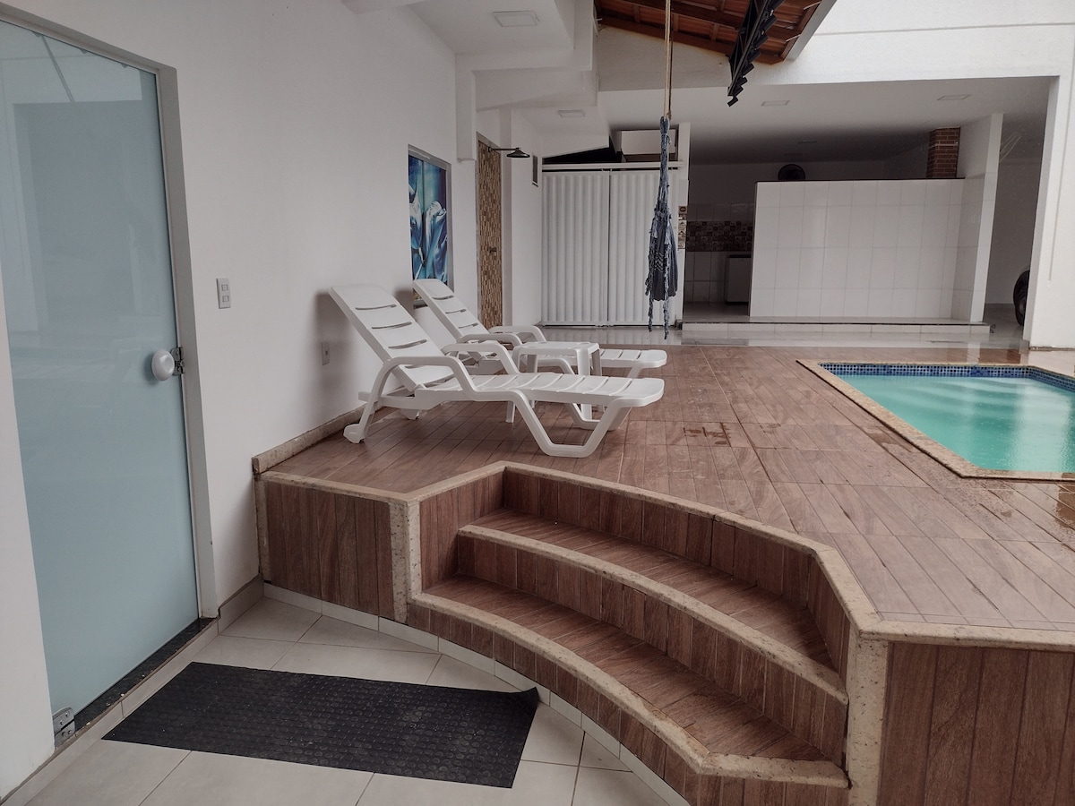 An inviting deck area features white lounge chairs positioned beside a small pool. The floor comprises wooden elements, and steps lead down to the pool area. A glass door overlooks the space, while an open kitchen is visible in the background.