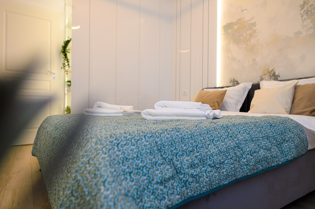 A spacious bed covered with a patterned blue throw is centrally positioned in the room. Crisp white towels are neatly folded at the foot of the bed. Soft lighting highlights the textured wall in the background, creating an inviting atmosphere.