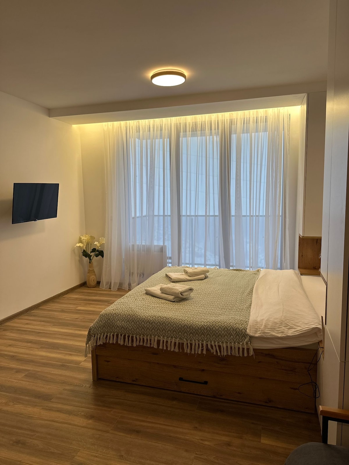 A modern bedroom features a spacious bed made with white linens and a green throw blanket. Large windows are dressed with sheer white curtains, allowing natural light to brighten the room. A flat-screen TV is mounted on the wall, complementing the warm wood accents.