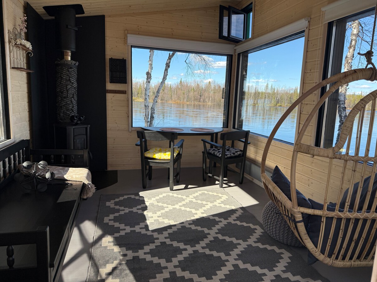 A light-filled seating area features large windows offering views of the serene water. Two black chairs are positioned around a small table, inviting relaxation. A woven hanging chair adds a comfortable touch, while a patterned rug enhances the cozy atmosphere.