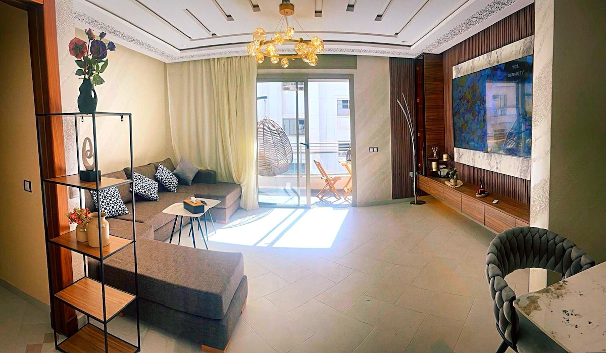 A bright living area is showcased, featuring a sectional sofa adorned with decorative cushions. A large window allows natural light to fill the space. A sleek entertainment unit is positioned against a wall, complemented by a minimalist shelf. A comfortable chair hangs in the corner.