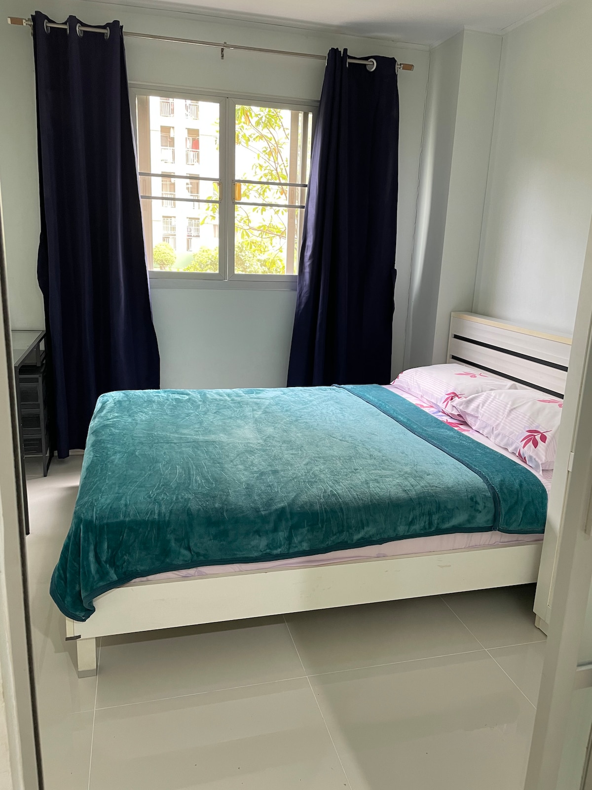 A comfortable bedroom features a spacious bed with a teal blanket draped over it. Natural light enters through a window, framed by navy blue curtains. A small desk is positioned nearby, and the room's floor displays a polished finish.