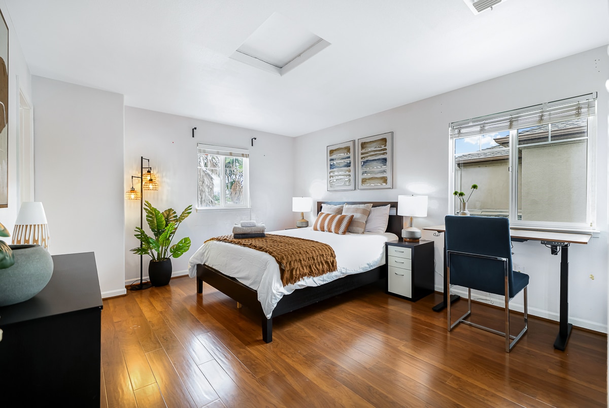A well-lit bedroom displays a spacious bed with neutral bedding, complemented by decorative throw pillows. A desk with a chair is positioned near a window, providing natural light. A potted plant adds a touch of greenery, and minimalist artwork adorns the walls.