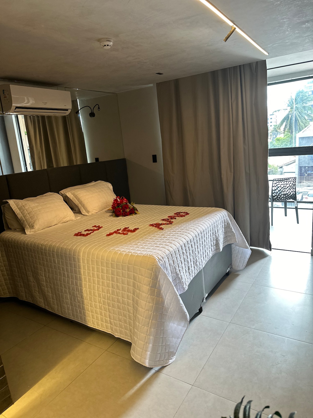 A spacious bedroom is highlighted by a neatly made bed covered with a white quilt. Decorative flowers are arranged on the bed. Natural light enters through large glass doors that lead to a small balcony, with curtains softly framing the view.