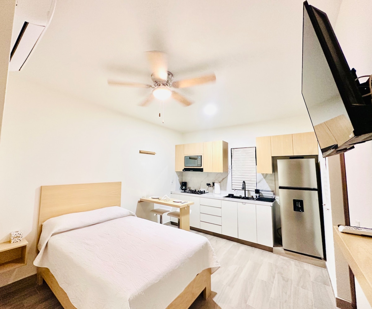 A well-organized studio layout features a spacious bed with a light-colored coverlet. A small dining area with a table is adjacent to a fully equipped kitchen. Dark cabinetry contrasts with light wood tones, while a ceiling fan and wall-mounted television contribute to a comfortable atmosphere.