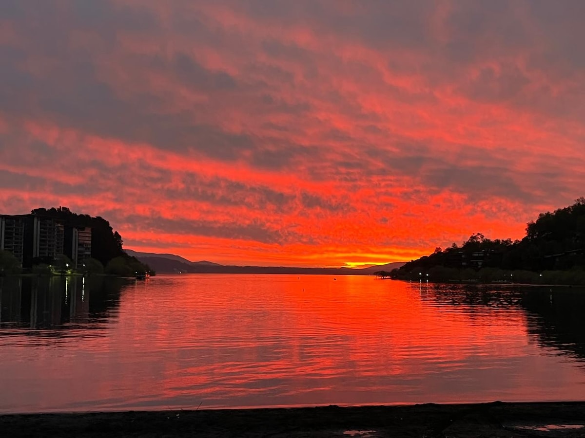 A vibrant sunset is reflected on the calm water, displaying shades of orange and red across the sky. The horizon features distant silhouettes of hills, while dark buildings line the shore, creating a contrast against the colorful backdrop.
