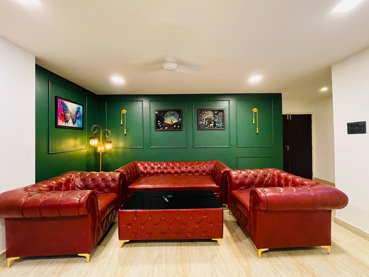 A spacious living area is highlighted by deep red leather sofas arranged around a sleek black coffee table. The green walls are adorned with colorful artwork, while soft lighting fixtures contribute to a relaxed ambiance.