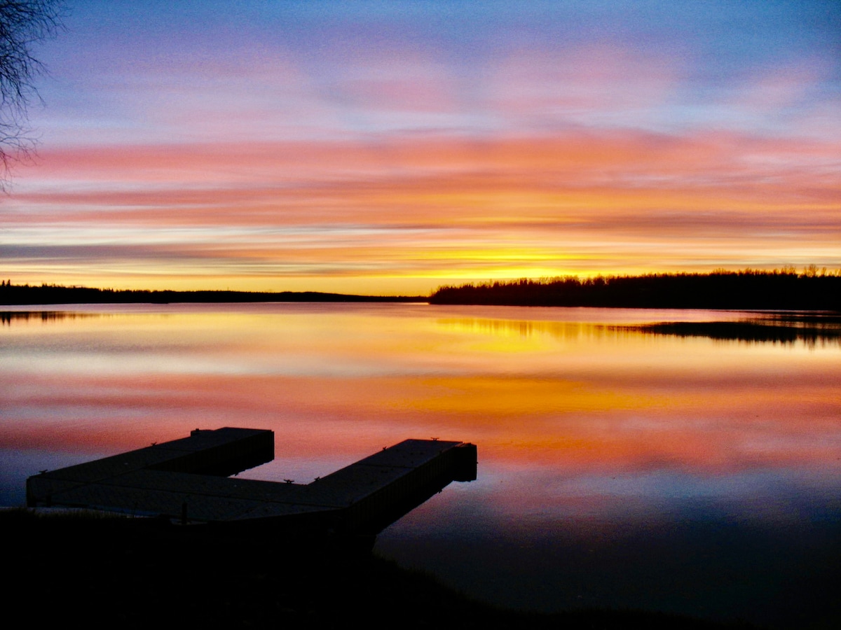 A serene lake scene is captured during sunset, displaying vivid hues of orange, pink, and purple reflecting off the water. A wooden dock extends into the lake, enhancing the tranquil atmosphere surrounded by distant trees silhouetted against the colorful sky.