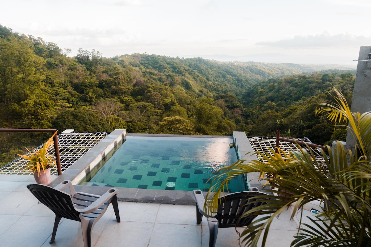 An infinity pool is situated on a terrace, offering unobstructed views of the lush green mountains in the distance. Two black lounge chairs are positioned beside the pool, enhancing the serene atmosphere. Potted plants add a touch of nature to the outdoor space.