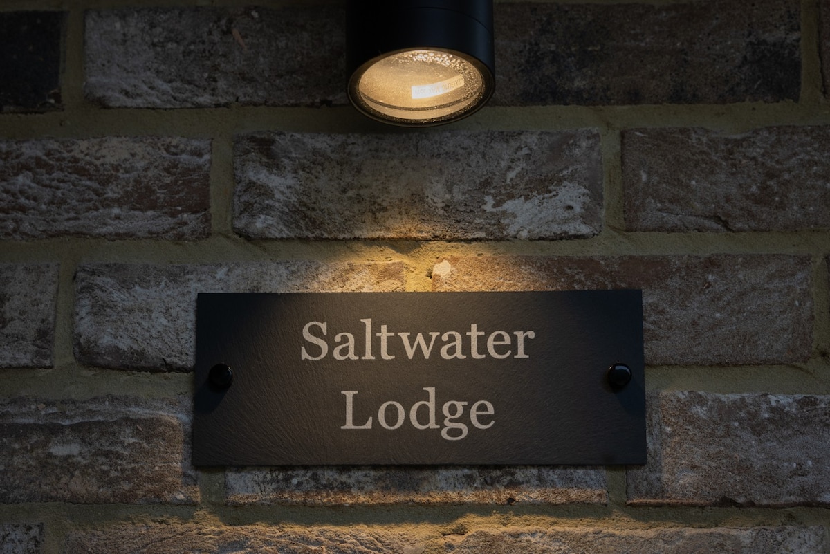 A sign labeled 'Saltwater Lodge' is mounted on a textured brick wall, subtly illuminated by a warm overhead light. The contrasting colors of the sign and bricks create a welcoming entrance feature.