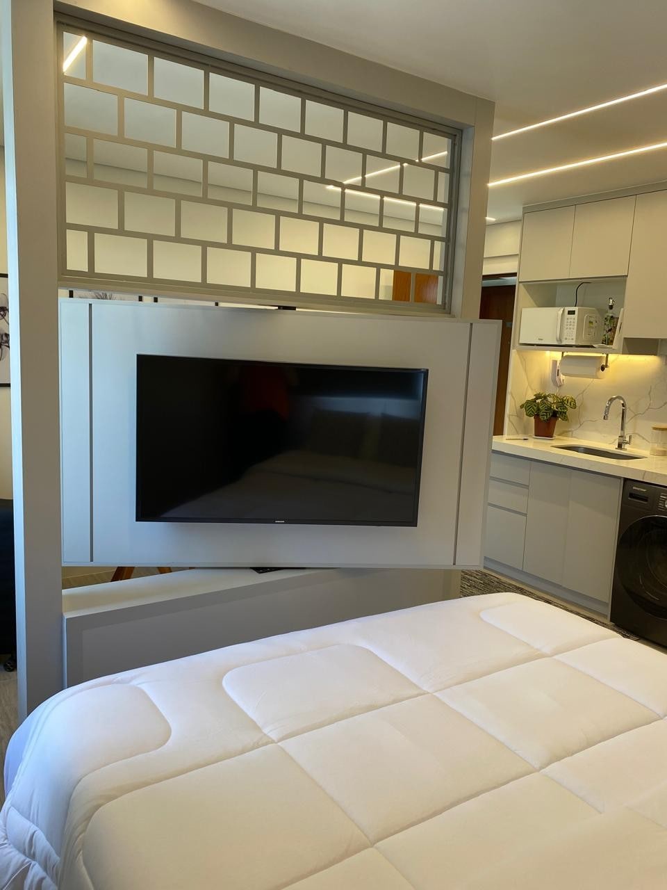 A modern bedroom features a soft bed with crisp white linens, positioned adjacent to a sleek wall-mounted television. The open-layout creates a seamless transition to a fully equipped kitchen, with minimalistic cabinetry and convenient appliances visible in the background.