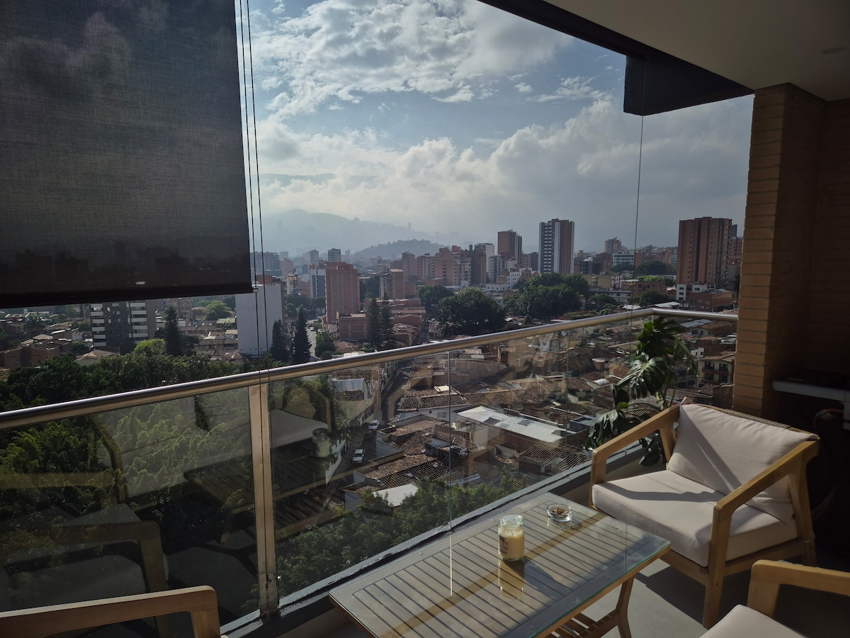 A balcony offers a comfortable seating arrangement with wooden chairs and a table. The view captures the city of Medellín, showcasing a mix of modern buildings and mountainous backdrop under a partly cloudy sky.