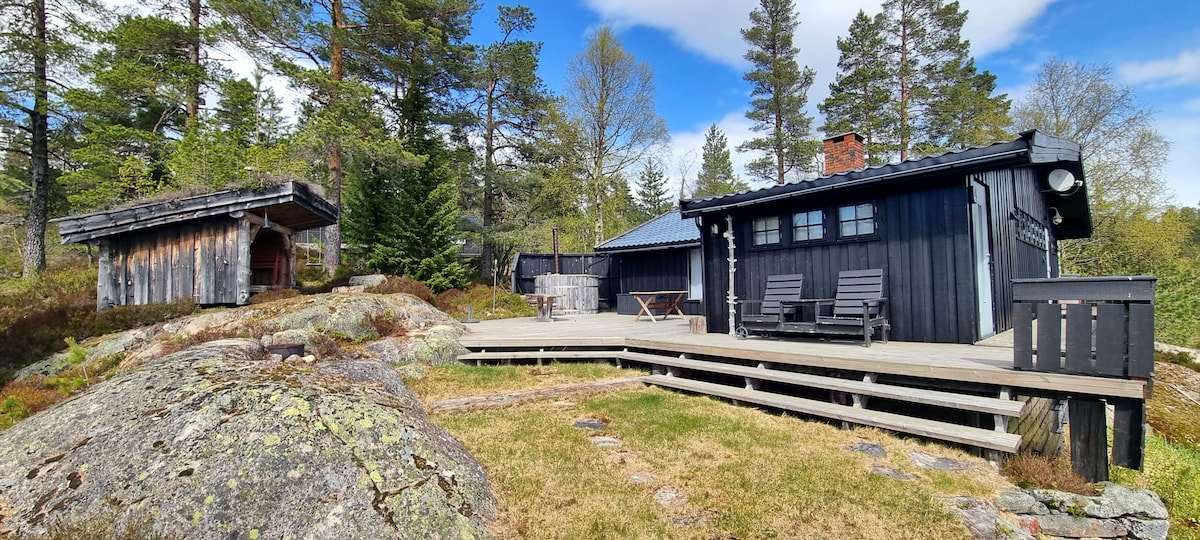 An outdoor view showcases a spacious wooden deck extending from the dark-hued cabin. A seating area with two chairs faces the surrounding pine trees, while a cozy shelter is positioned nearby. The natural landscape features rocky terrain and a mix of greenery.