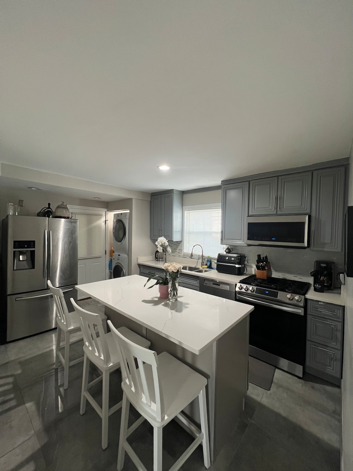 The kitchen features modern appliances, including a stainless steel refrigerator and oven. A central island with seating for three is complemented by white cabinetry. Natural light enters through a window above the sink, enhancing the workspace's functionality.