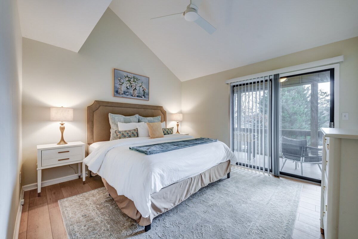 A spacious bedroom features a large bed with decorative pillows and a soft throw at the foot. A bedside table with a lamp is present on one side, while glass sliding doors open to a balcony with seating. Light wood flooring and neutral walls enhance the peaceful ambiance.