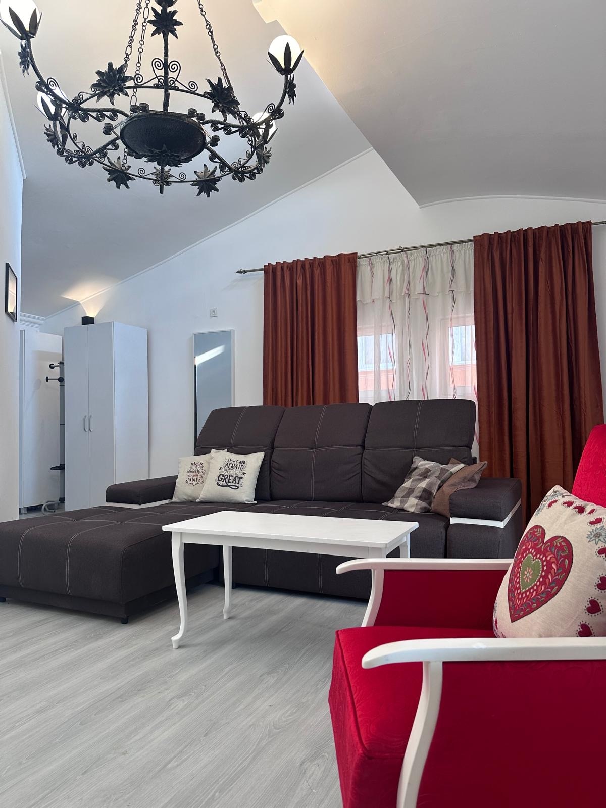 A spacious living area is highlighted by a dark sectional sofa paired with a coordinating ottoman. A white coffee table is positioned in front. A red accent chair adds a pop of color, while large windows with sheer curtains allow natural light to enhance the room.