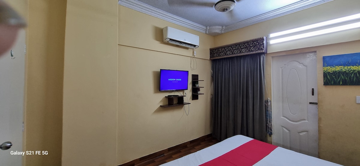 The bedroom features a spacious bed with a red accent on the bedding. A flat-screen TV is mounted on the wall opposite a window, which is covered by a gray curtain. An air conditioning unit is positioned above, contributing to the room's comfort.