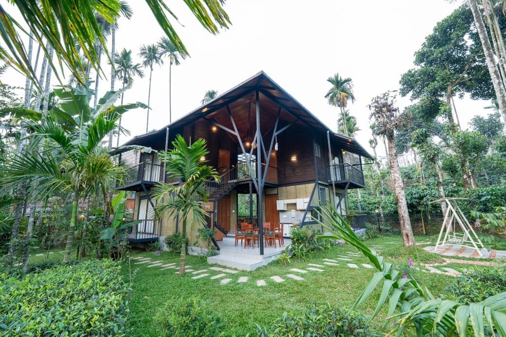 A two-story house is framed by palm and other tropical plants. The structure features a modern design with large windows and a wooden balcony. The surrounding area is landscaped with manicured grass and pathways leading to the entrance, creating a harmonious connection with nature.