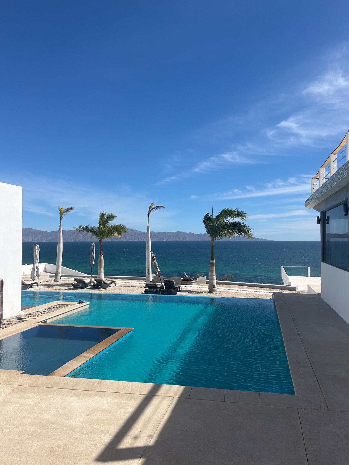 A large outdoor pool is positioned against a backdrop of the sea, with palm trees gently swaying in the breeze. Loungers line the poolside, and distant mountains are visible across the water under a clear blue sky.