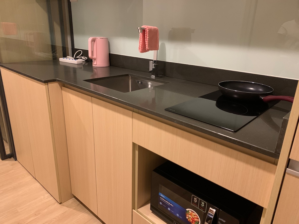 The kitchen area is equipped with a sleek countertop featuring a sink and an induction cooktop. A pink kettle adds a pop of color, while a microwave is tucked neatly beneath the counter for convenience. A dishcloth hangs nearby for easy access.