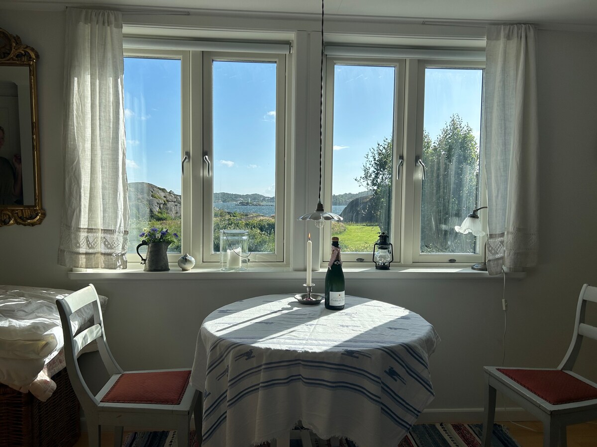 A dining area is illuminated by natural light from large windows, offering a view of the surrounding landscape. A round table with a white tablecloth is set with candles, accompanied by two chairs. Decorative plants and lanterns enhance the inviting atmosphere.