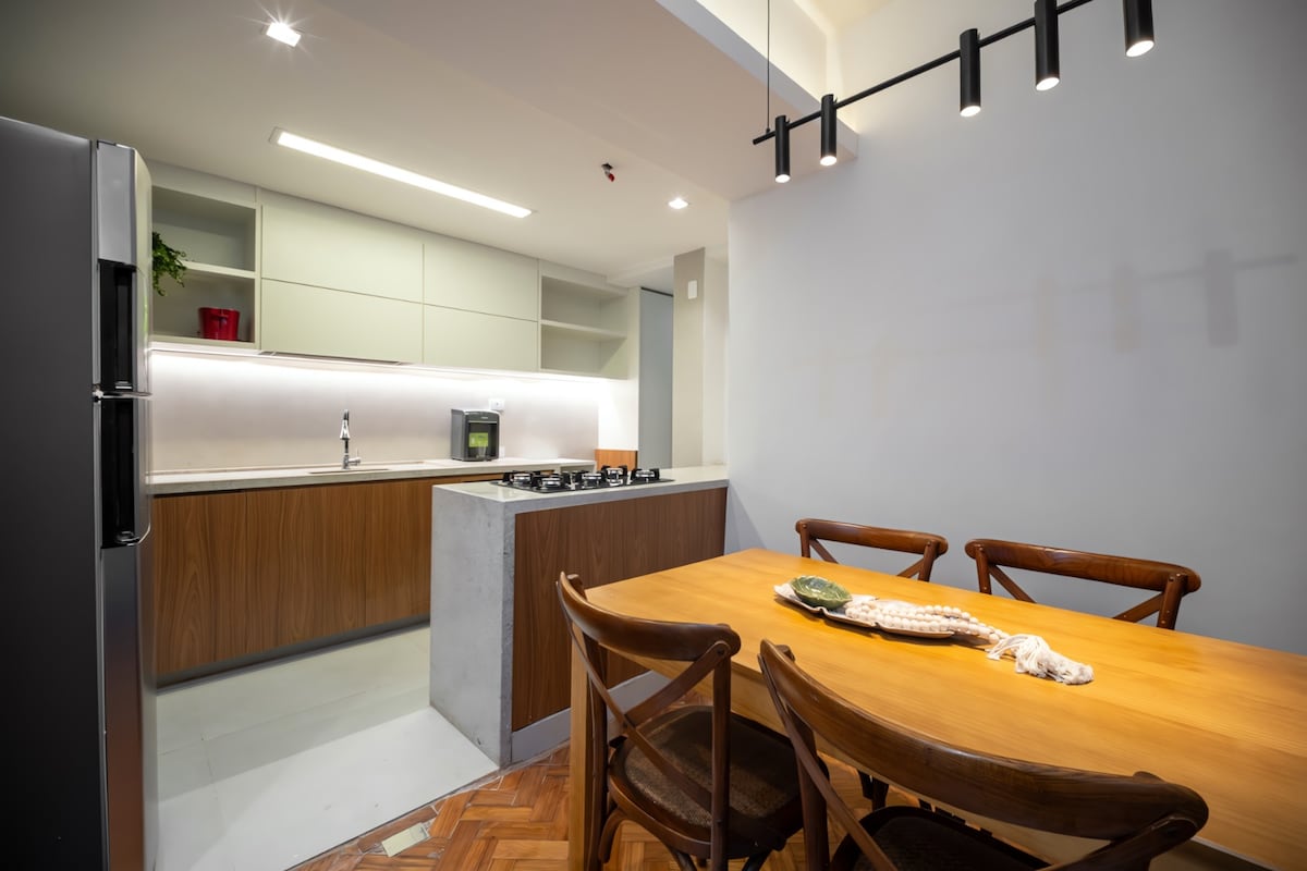 A modern kitchen is shown, featuring sleek cabinetry and a spacious countertop. A dining table with wooden chairs is positioned in the foreground, adorned with a decorative centerpiece. Bright lighting illuminates the area, enhancing the clean design and functionality.