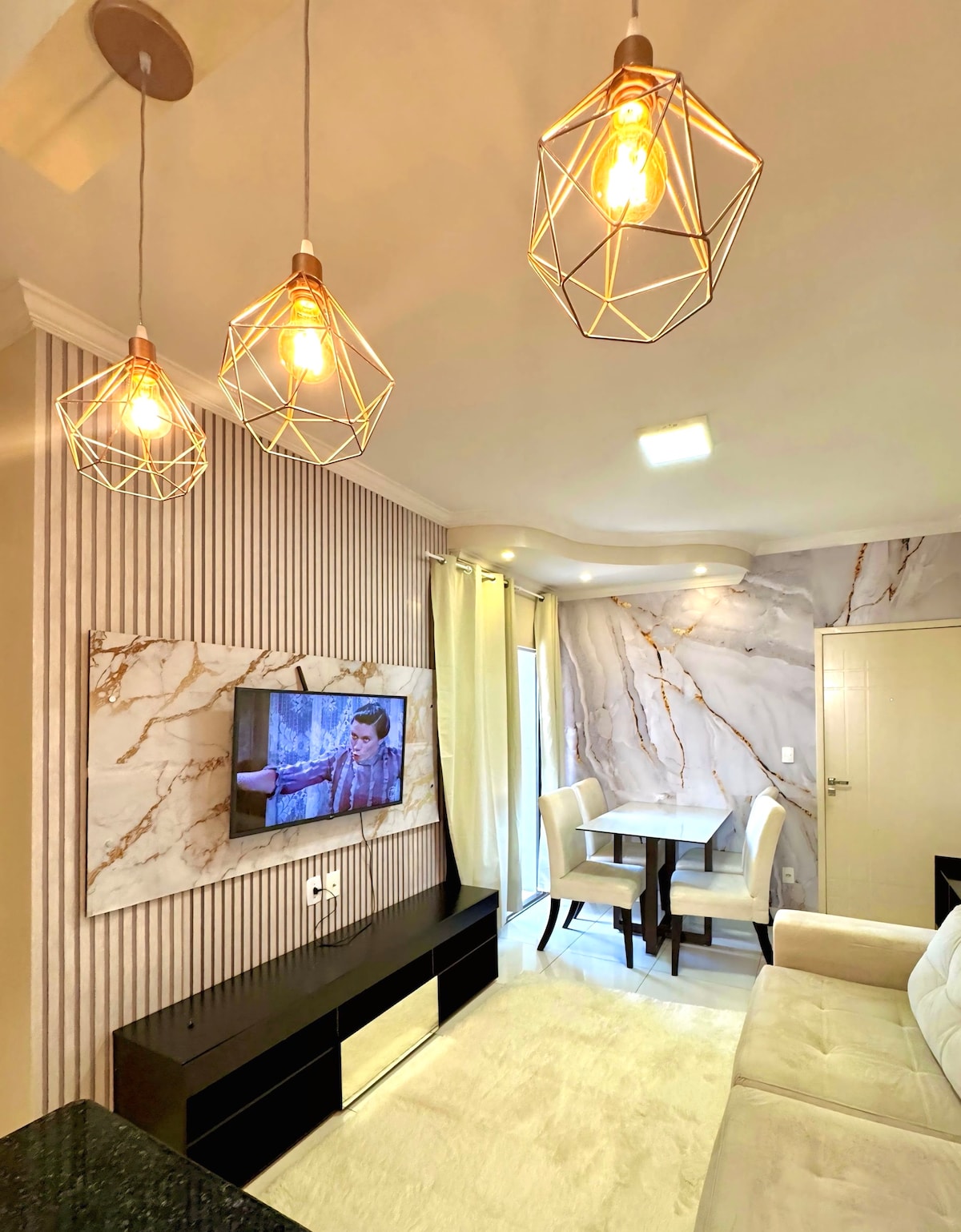 A modern living area showcases a sleek television mounted on a decorative wall featuring marble-like designs. Stylish pendant lights hang above a cozy seating arrangement, while a dining table with two chairs is positioned nearby, creating an open and inviting space.
