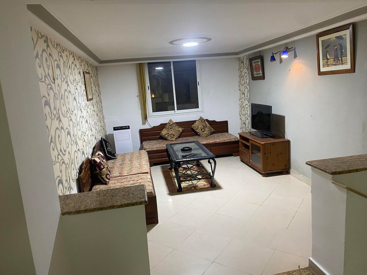 A cozy living room features a comfortable seating area with patterned cushions on wooden benches. A central coffee table is positioned in front of a TV, with a window allowing natural light. Decorative wall art enhances the space's character.