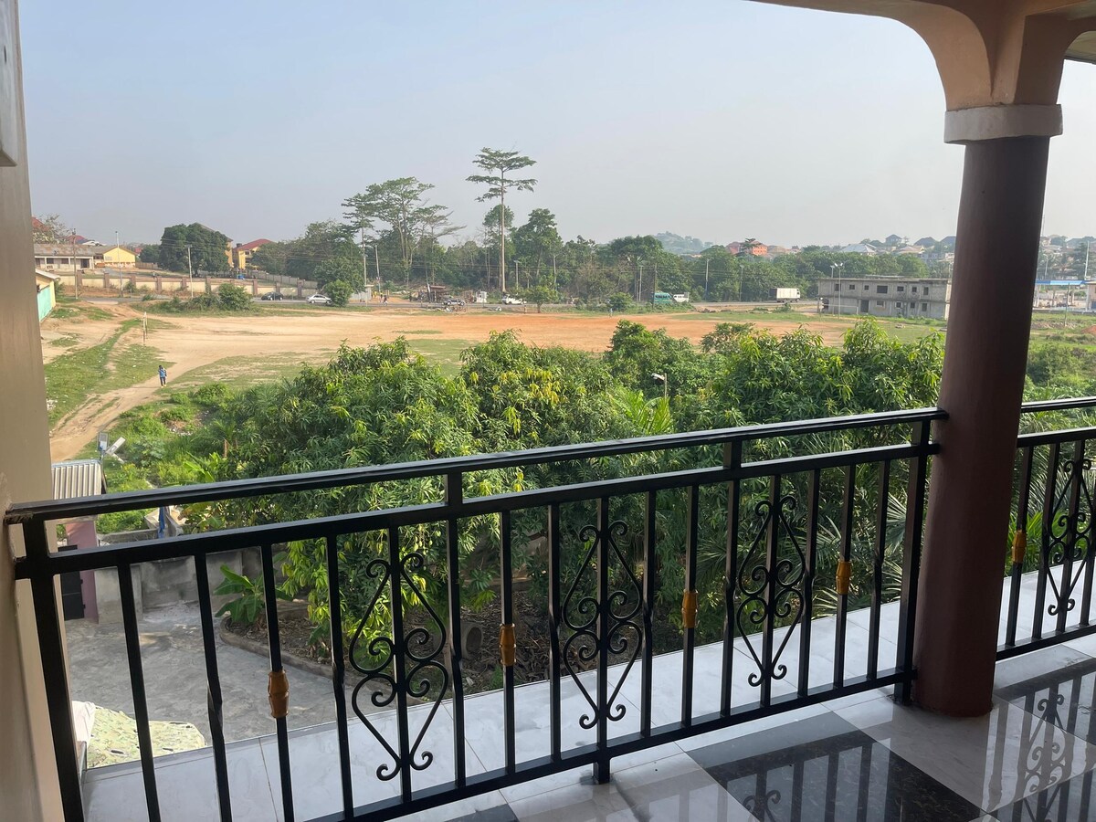 A balcony view overlooks a lush green landscape. The expansive field is bordered by trees and distant buildings under a clear sky. A black wrought iron railing complements the tiled flooring of the balcony, providing a spacious area for enjoying the outdoors.