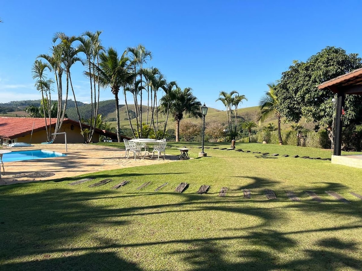 An outdoor area features a refreshing pool surrounded by well-maintained grass and tropical plants. White outdoor furniture is visible, including a dining set. A path of stepping stones leads through the manicured lawn, under a clear blue sky.