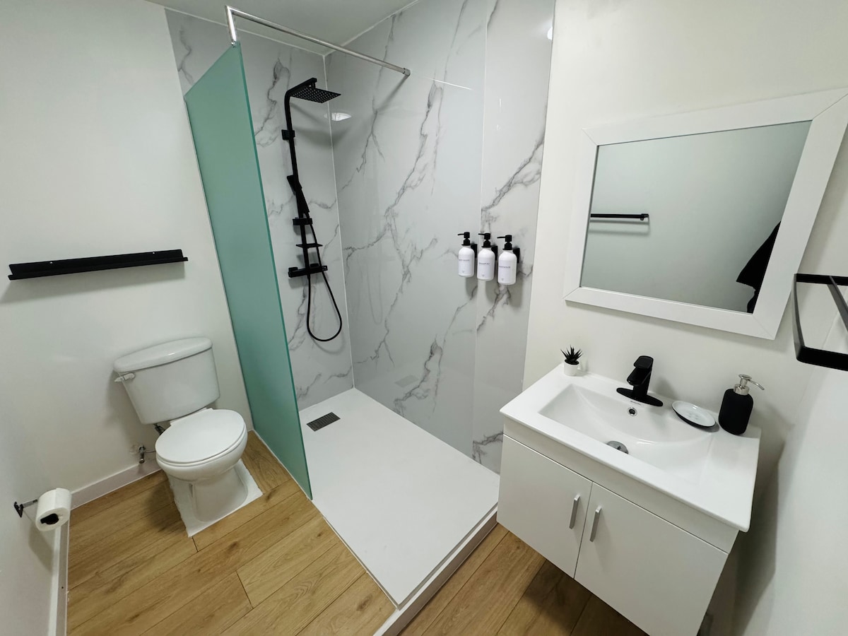 A modern bathroom features a spacious walk-in shower with a rainfall showerhead and marble-inspired walls. A clean white vanity with a large mirror complements the contemporary black fixtures. Essential toiletries are neatly arranged, and natural light enhances the bright ambiance. A minimalist shelf adds practical storage.