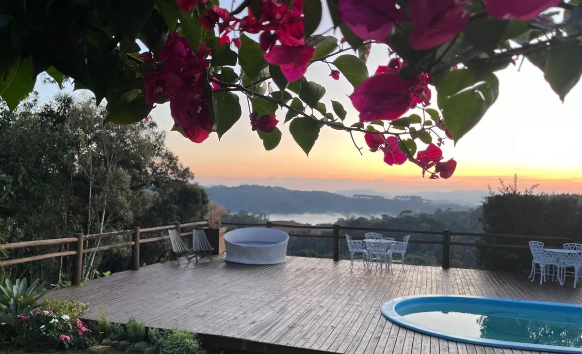 A wooden deck is adorned with vibrant pink flowers, framing a tranquil view of the sunrise over rolling hills. A circular hot tub and a swimming pool invite relaxation, while white outdoor furniture is arranged for seating. Lush greenery surrounds the area, enhancing the natural beauty.