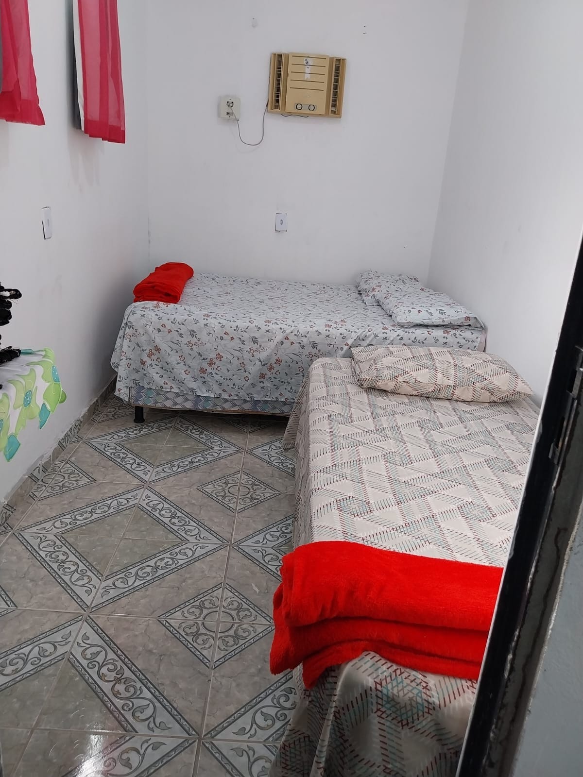 A small bedroom contains two beds dressed in floral and patterned bed linens. Red towels are neatly placed on each bed. The tiled floor features a geometric design, and sunlight filters through sheer red curtains, enhancing the bright and airy feel of the room.