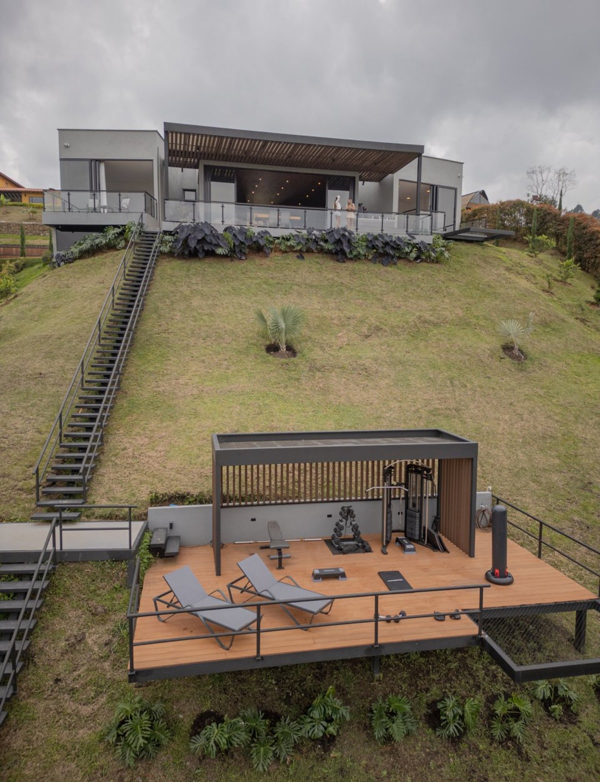 A modern house is situated on a hillside, featuring multiple levels and expansive glass sections. Below, a fitness area is displayed on a wooden deck, complete with exercise equipment and lounge chairs. A grassy landscape surrounds the property, enhancing its connection to nature.