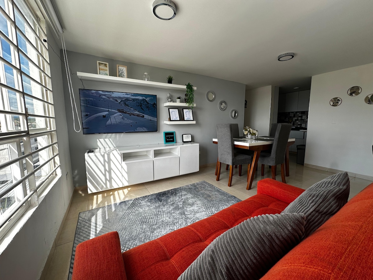 The living area features a vibrant red sofa, positioned adjacent to a modern entertainment unit with a large wall-mounted TV. A dining table can be seen in the background, complemented by stylish decor on the walls and natural light streaming through the spacious windows.