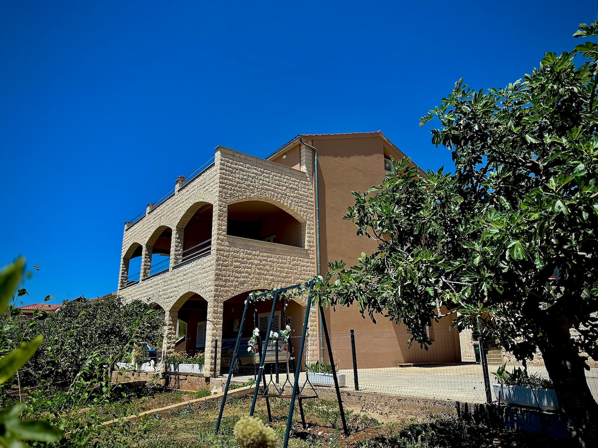 The large, two-story building features a textured stone façade and multiple balconies. A vibrant blue sky serves as a backdrop, while swings are visible in the well-kept yard, indicating a family-friendly atmosphere. Surrounding greenery adds to the outdoor space's charm.