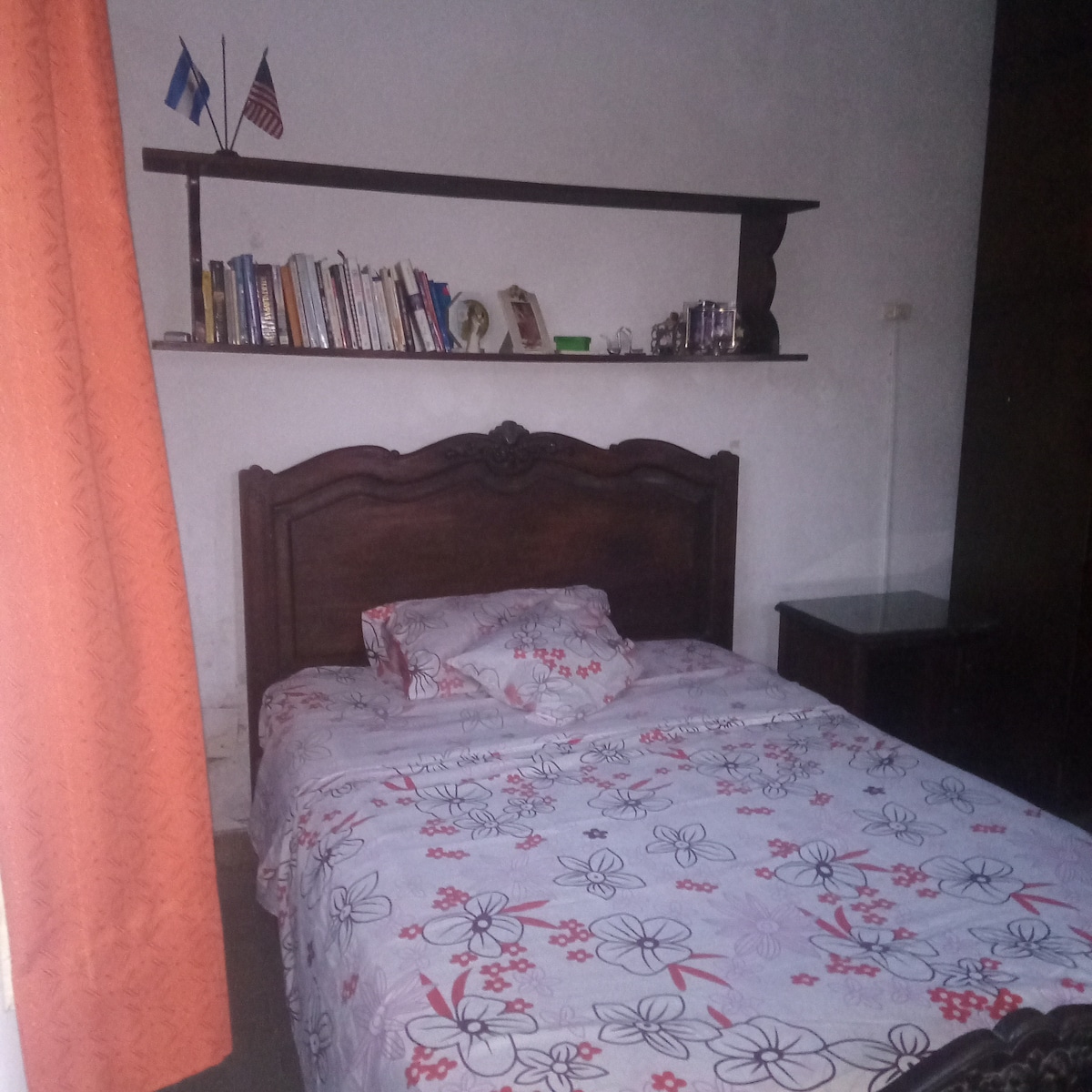 A cozy bedroom featuring a wooden headboard and a floral-patterned bedspread. A nightstand is positioned beside the bed. A shelf above the bed holds books and decorative items, while two flags are displayed on the wall, adding a touch of character to the room.