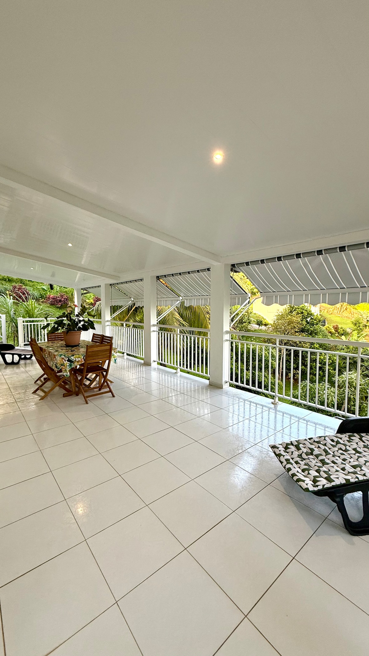 A spacious terrace is presented, featuring a dining table surrounded by wooden chairs. The floor is tiled in white, reflecting natural light. Lush greenery is visible beyond the railing, enhancing the serene atmosphere of the outdoor space.